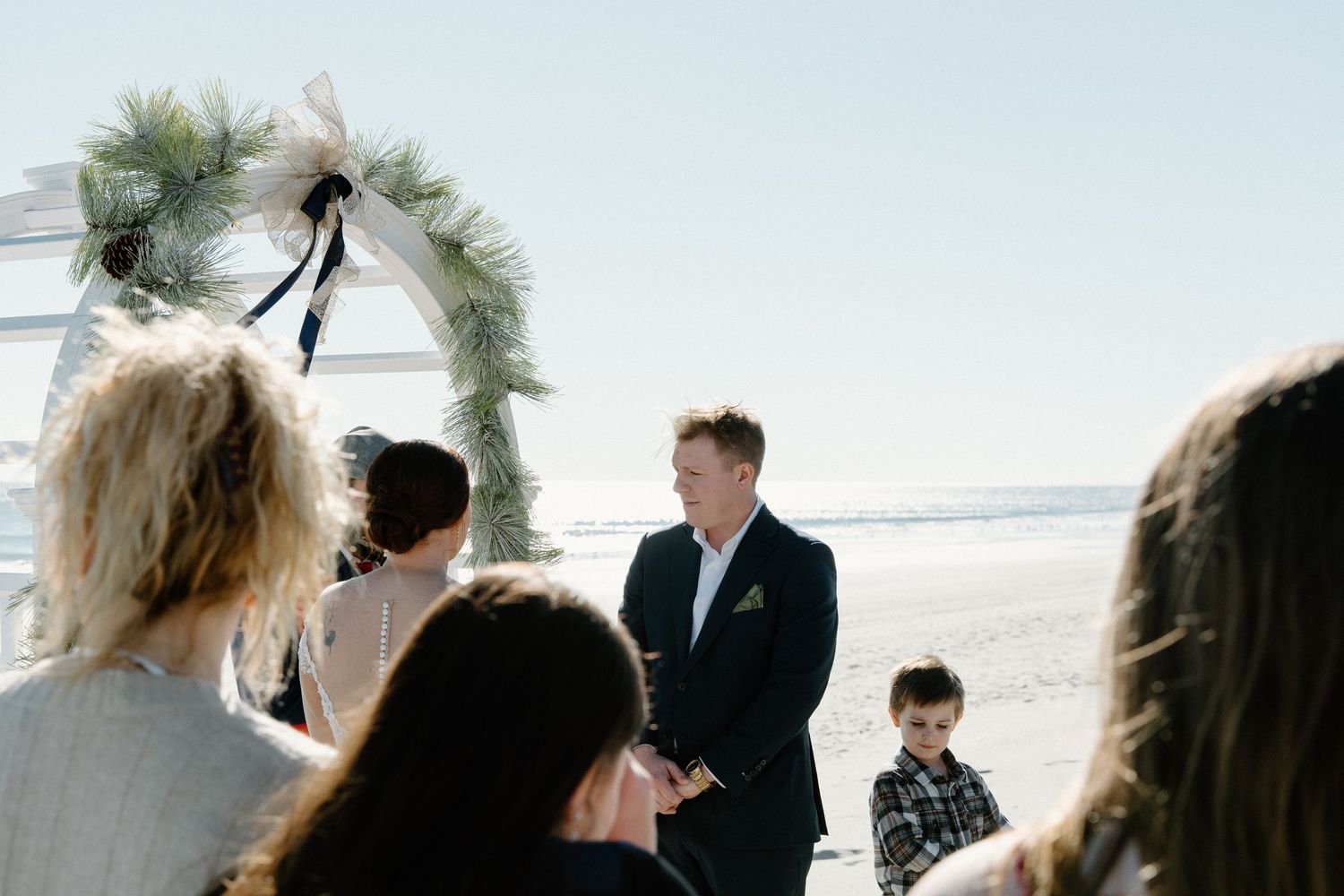 An outdoor beach wedding ceremony with guests gathered around a decorative floral arch by the ocean.