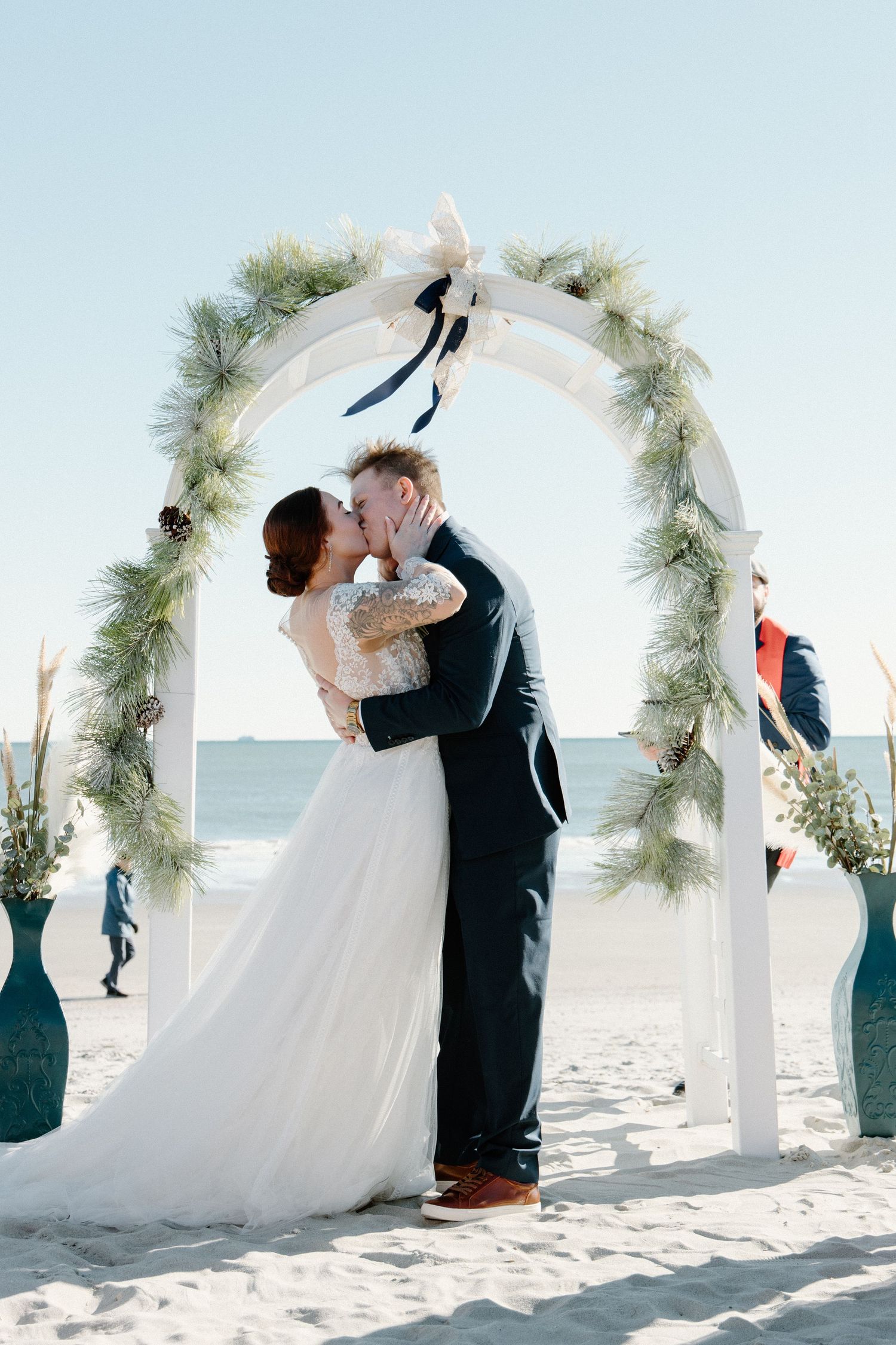 Couple shares romantic kiss under white floral arch during beach wedding ceremony with ocean backdrop.