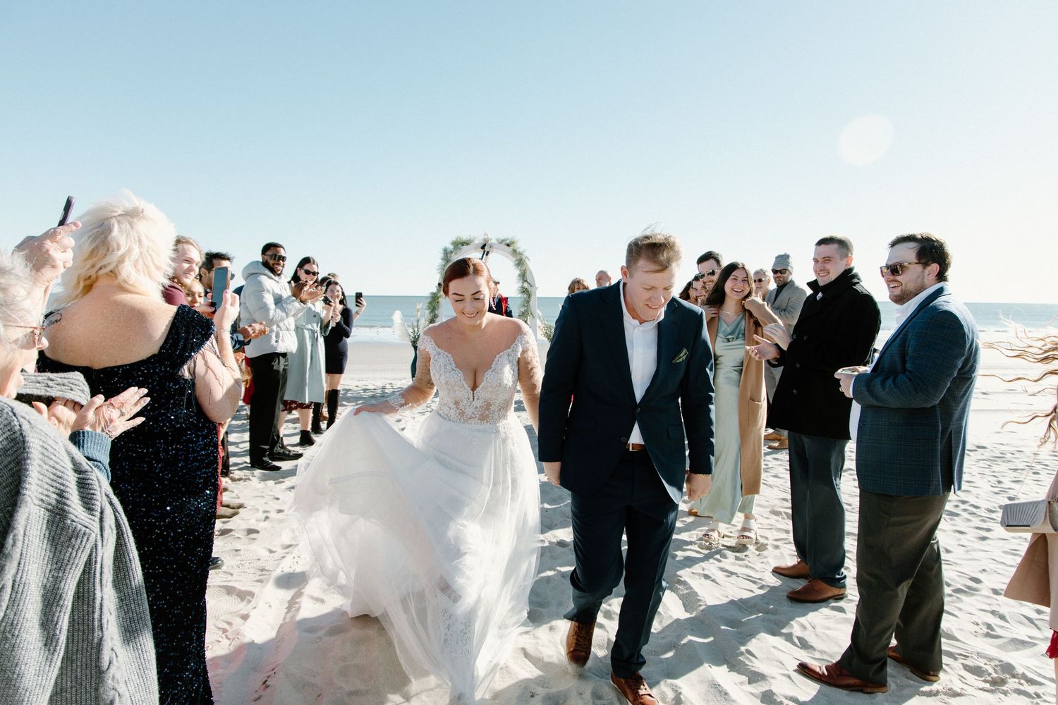 Newlyweds walk down aisle on sunny beach as wedding guests toss petals and celebrate their joyful ceremony.