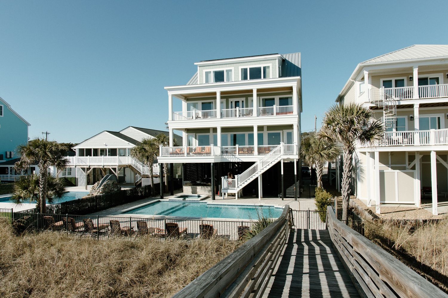 Beachfront homes with walkways leading to the sand stand elevated on stilts along a coastal shoreline during sunset.