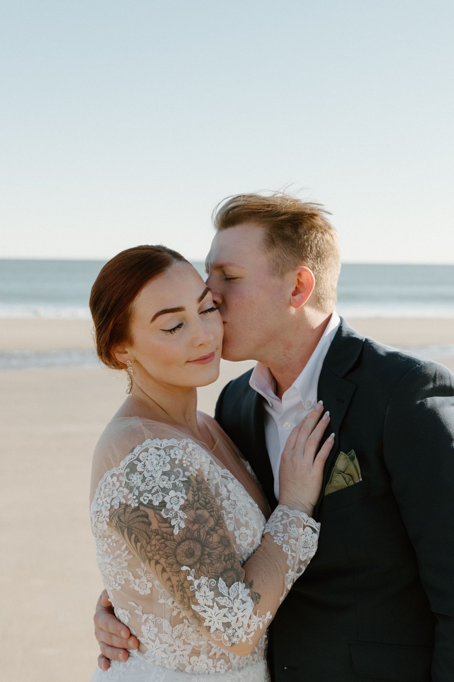 Romantic couple embraces on beach at sunset, with bride wearing lace wedding dress and groom in dark suit.