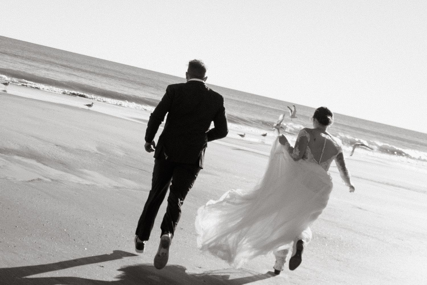 A black and white photo of a couple running joyfully on a beach, with the bride's wedding dress flowing in the wind.