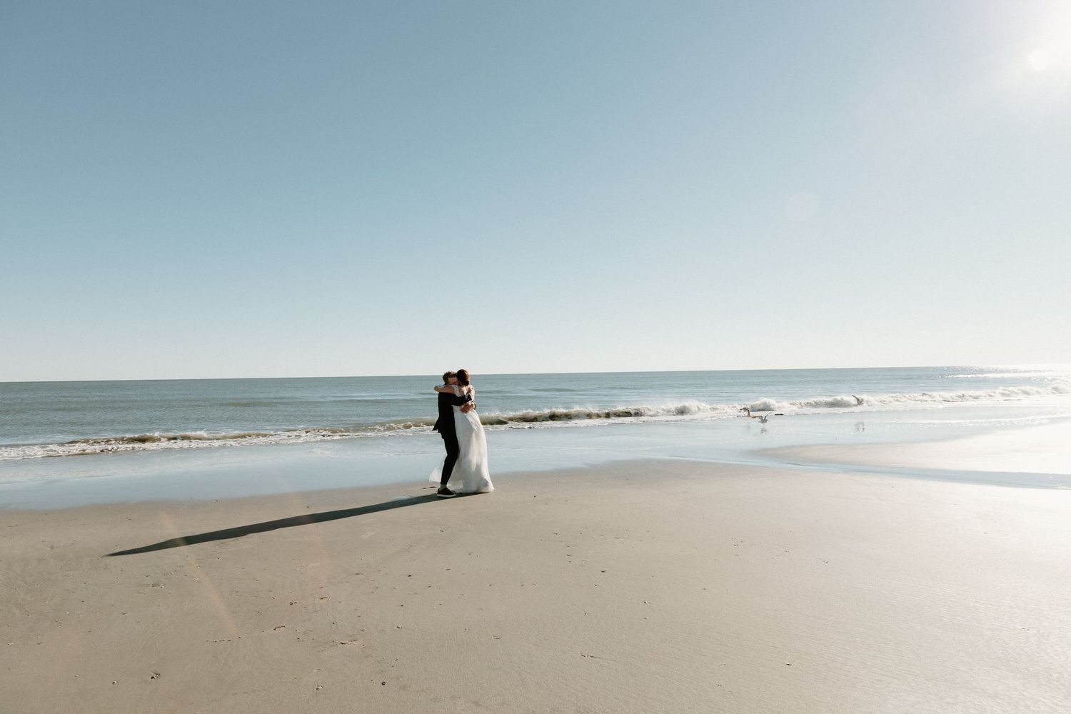 A couple embraces on a sunny, empty beach with waves rolling onto the shore and bright sunlit sky overhead.