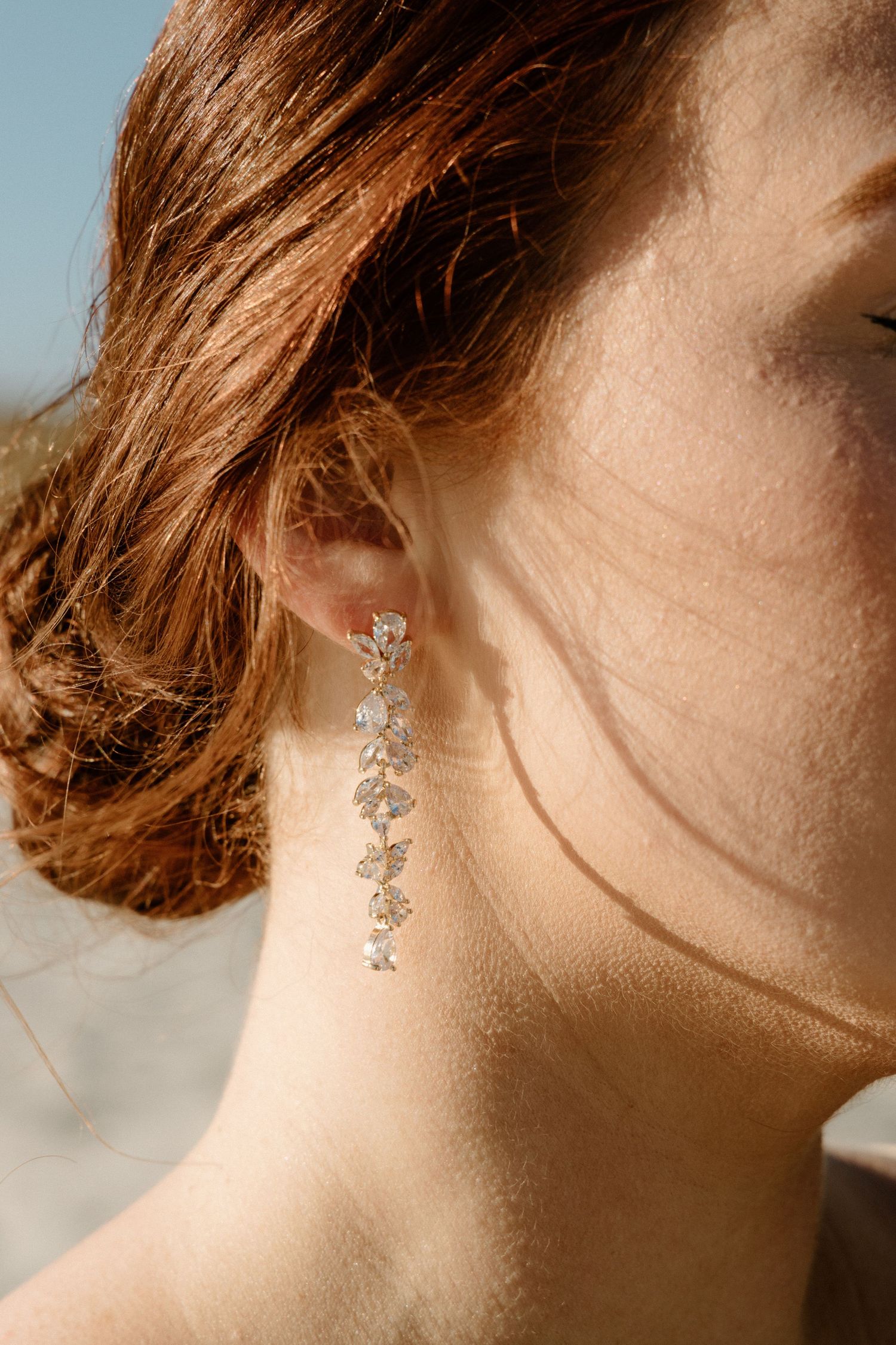 Delicate crystal dangle earring sparkles against auburn hair and soft neck in warm sunlight.