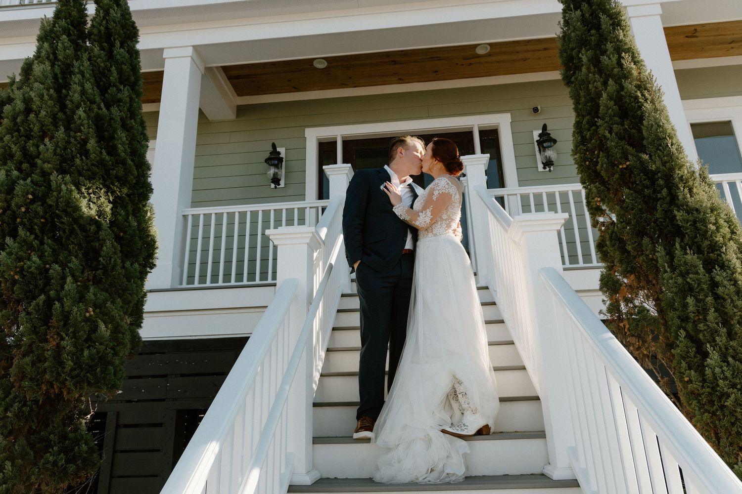 A couple embraces on an elegant white porch staircase flanked by tall cypress trees at a classic southern home.