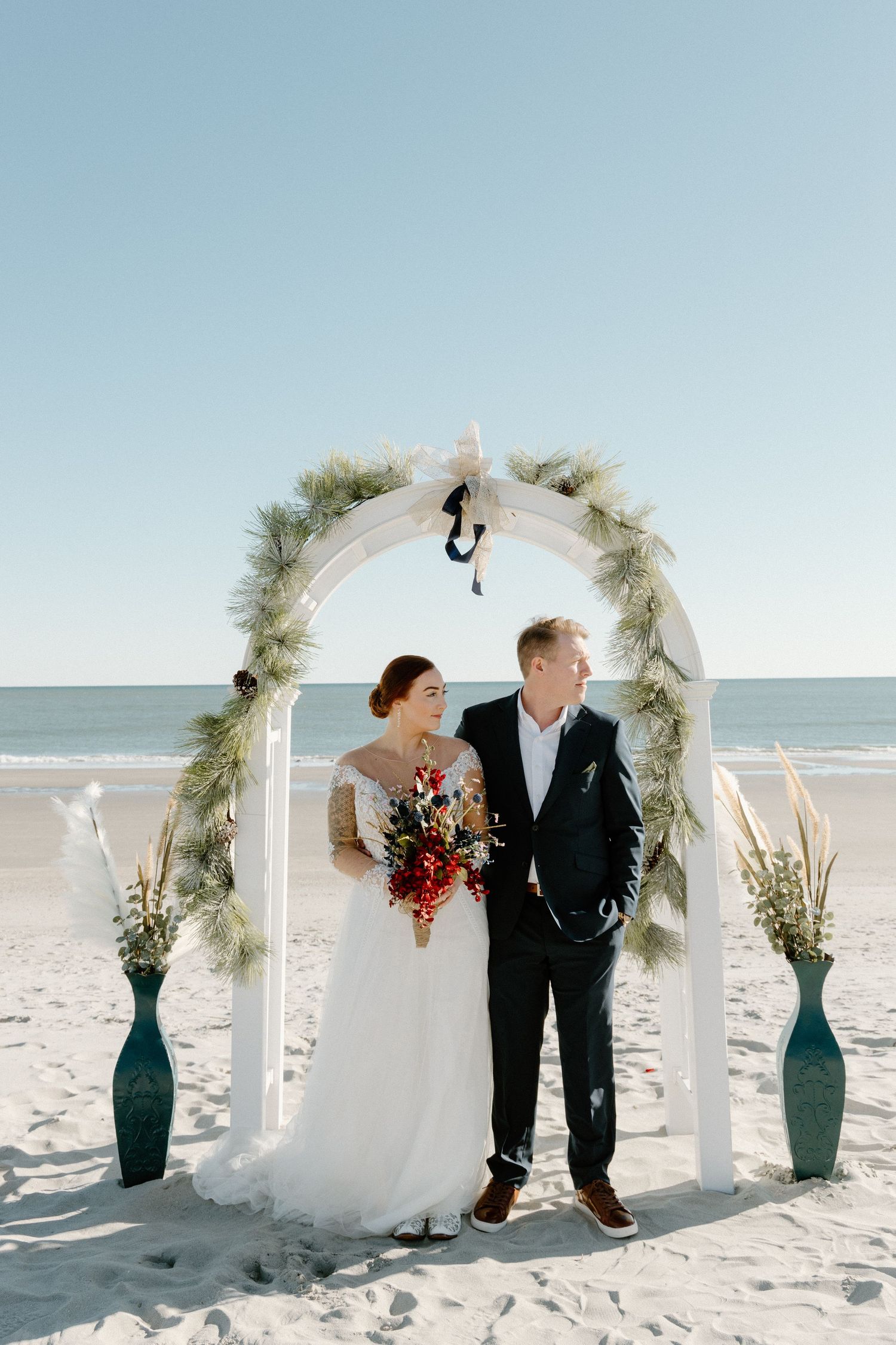 A couple stands under a white floral arch during their beach wedding ceremony with teal vases and ocean backdrop.