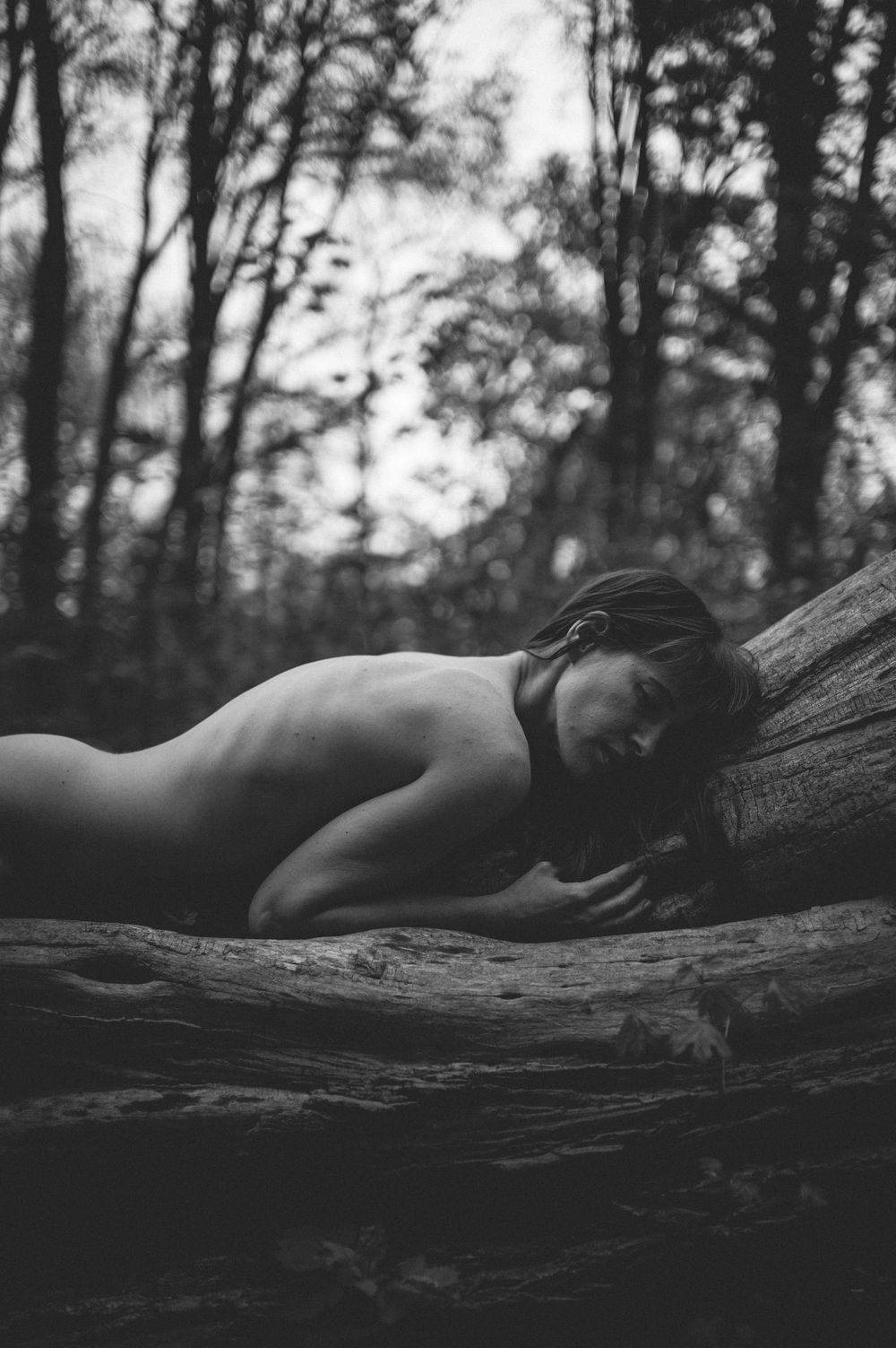 Black and white artistic portrait of a reclining figure on a fallen tree in a moody forest setting.