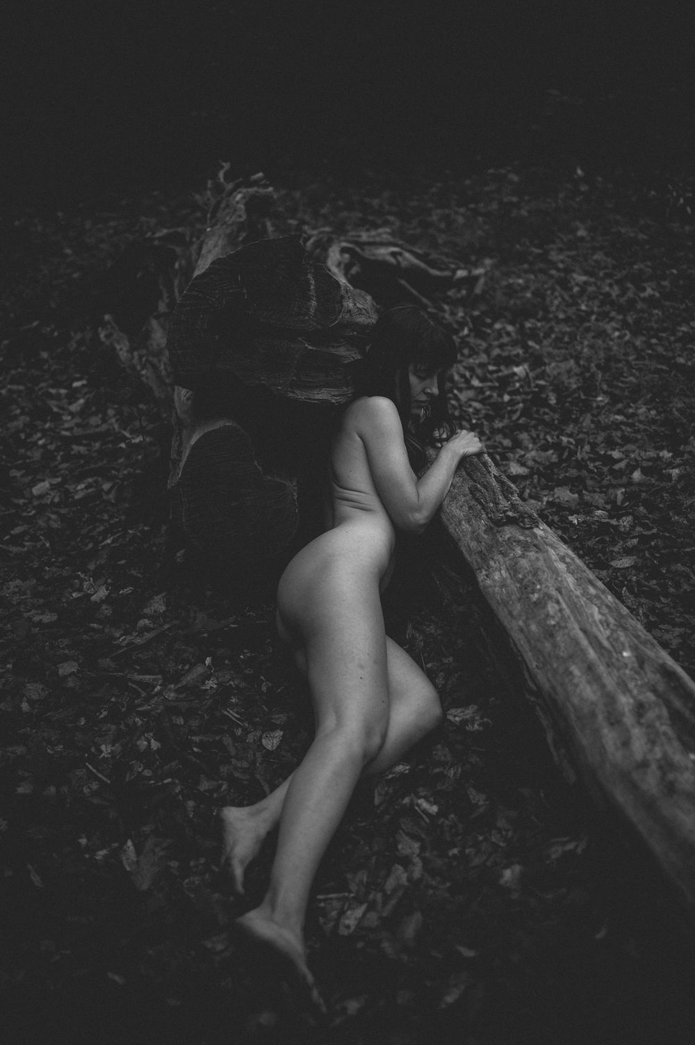 A black and white artistic nude photograph shows a figure reclining on a fallen log in nature.
