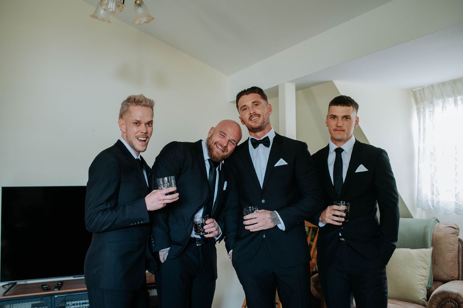 Four groomsmen in dark suits smiling and holding drinks while celebrating in a bright living room interior.