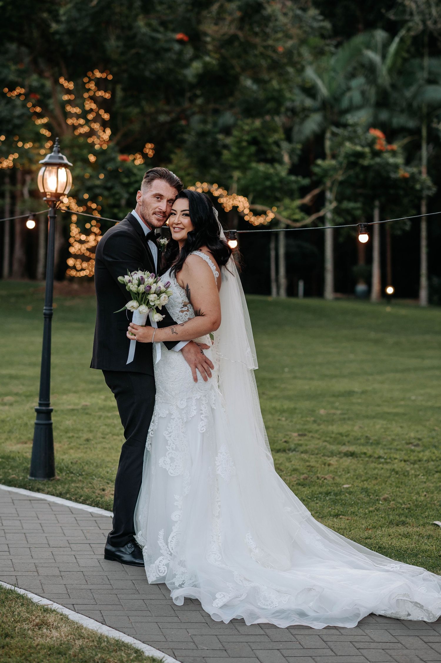A bride in a white lace gown and groom in black suit embrace on a lit garden path at dusk with string lights overhead.