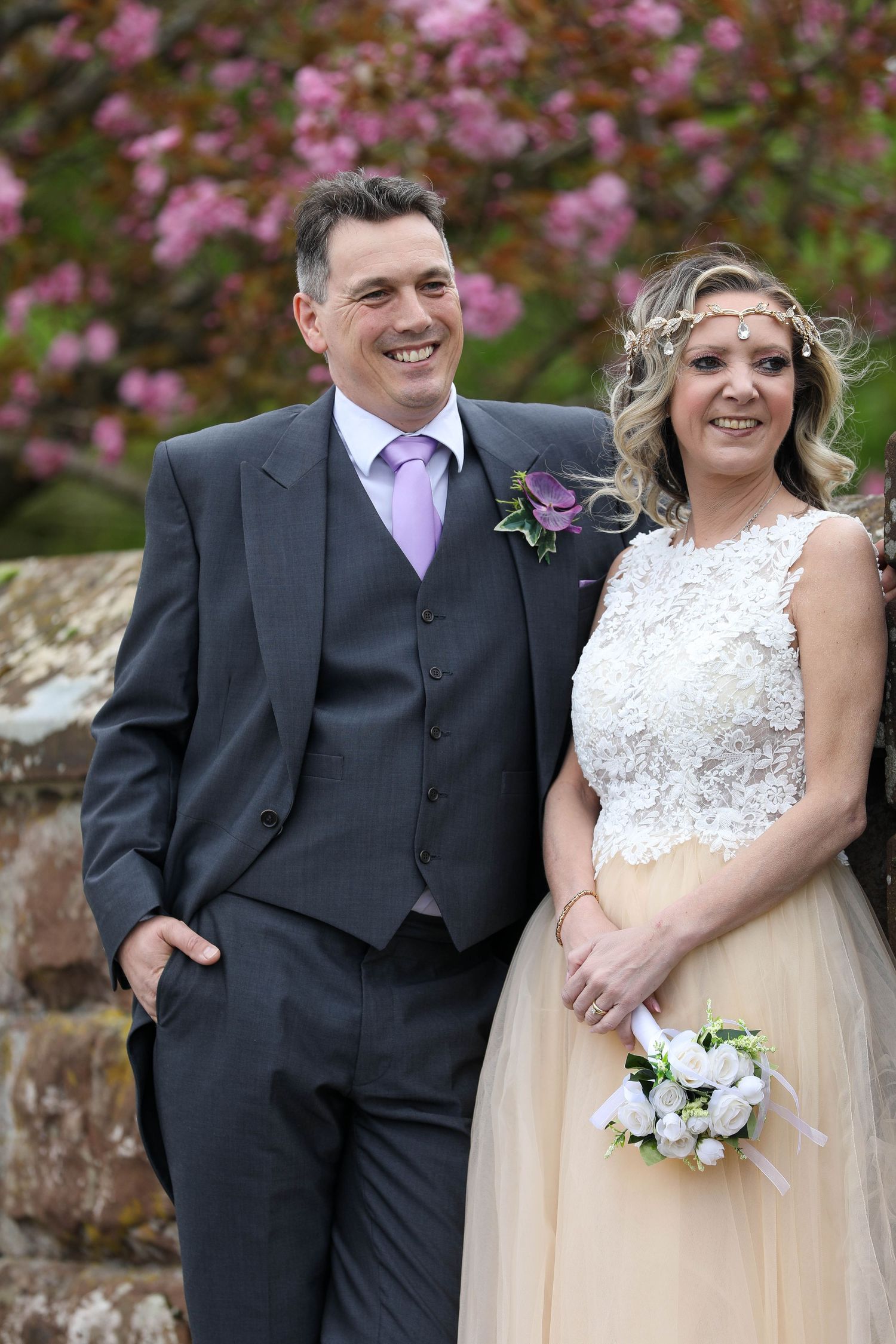 Happy newlyweds pose together in formal attire against blooming spring flowers.