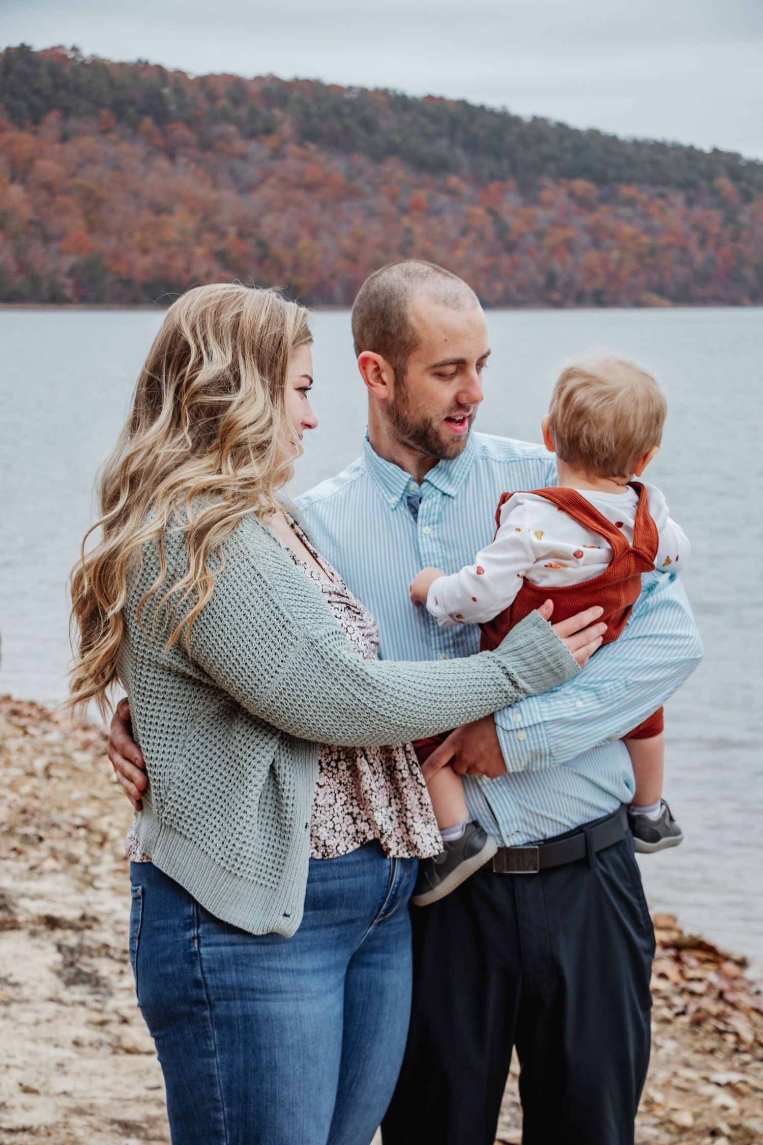 Smiling parents holding their toddler at a lakeside with colorful autumn foliage in the background.
