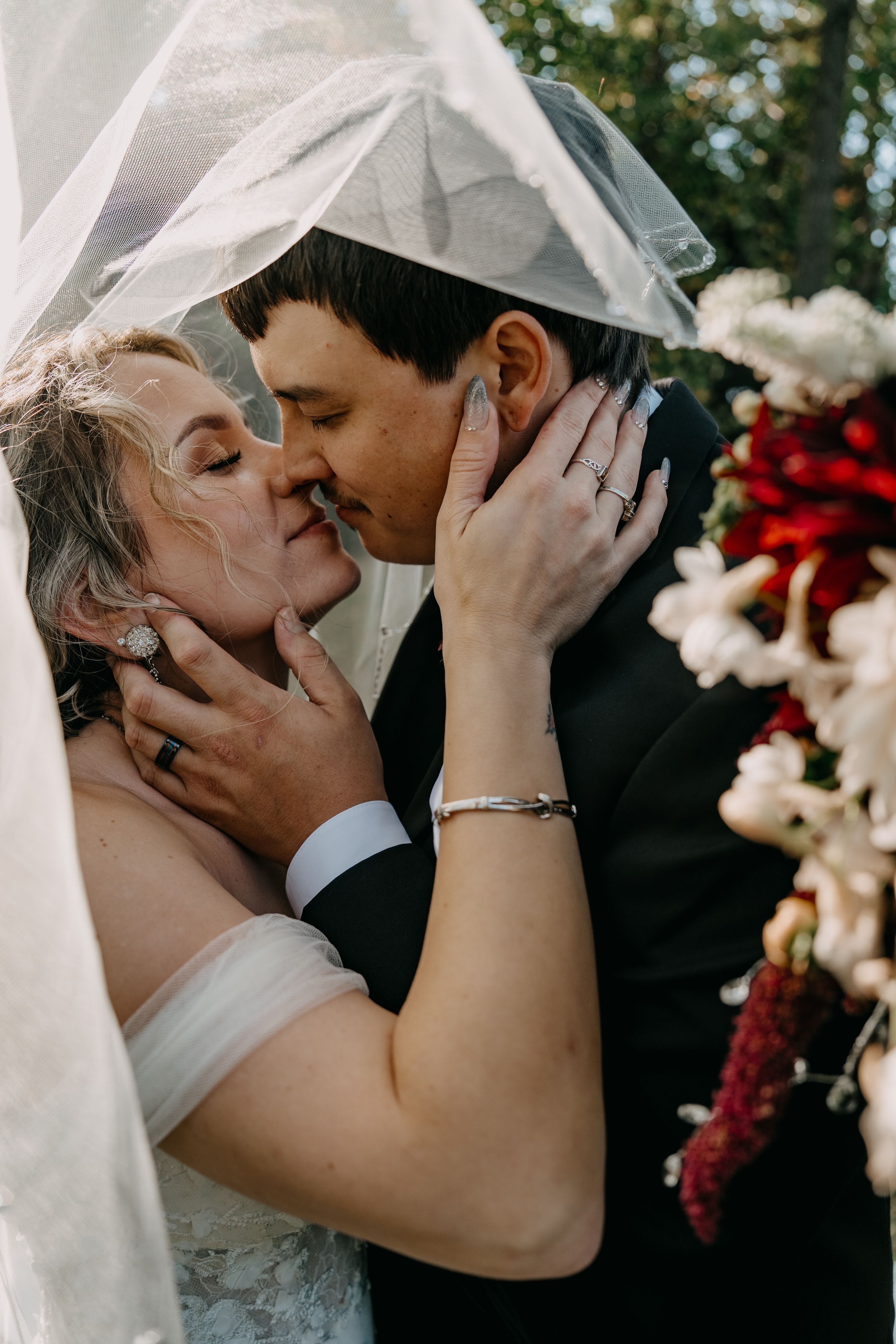 A romantic wedding photo of newlyweds sharing a tender kiss under a veil, surrounded by red and white floral decorations.