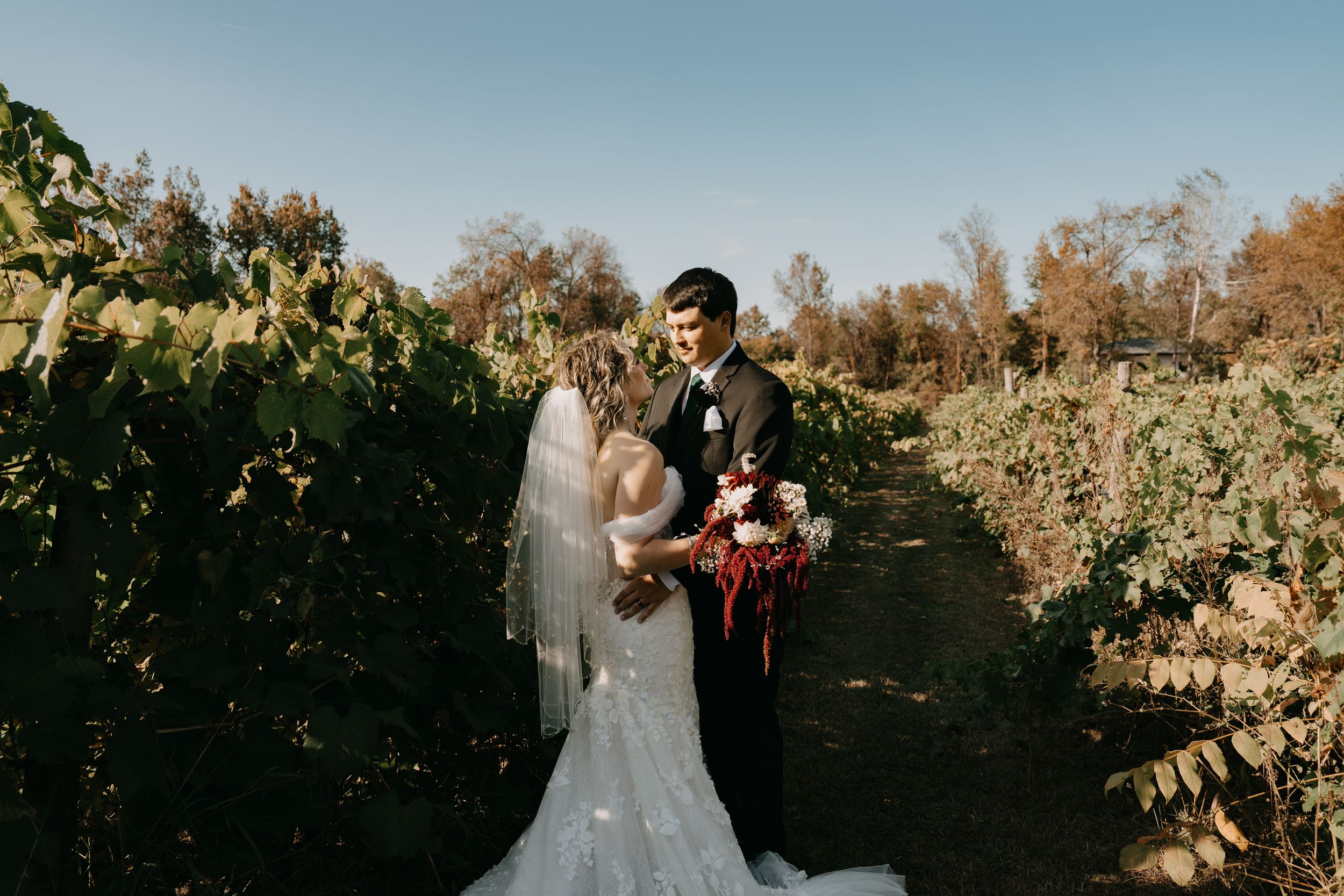 A couple shares a romantic moment in a sunlit vineyard with fall foliage and a beautiful floral bouquet.