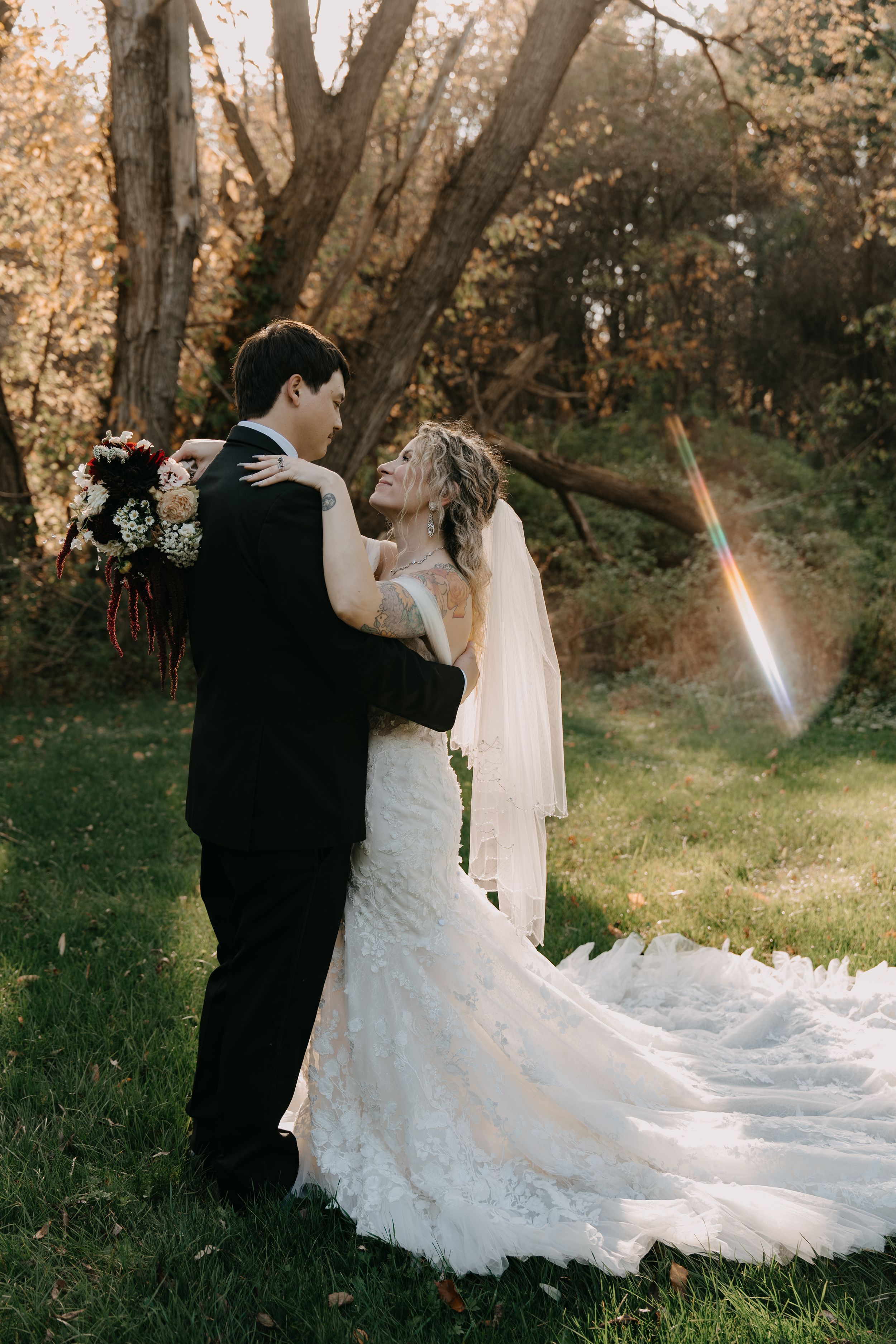 A couple in wedding attire embraces outdoors in a sunlit forest setting with a flowing white lace gown and veil.