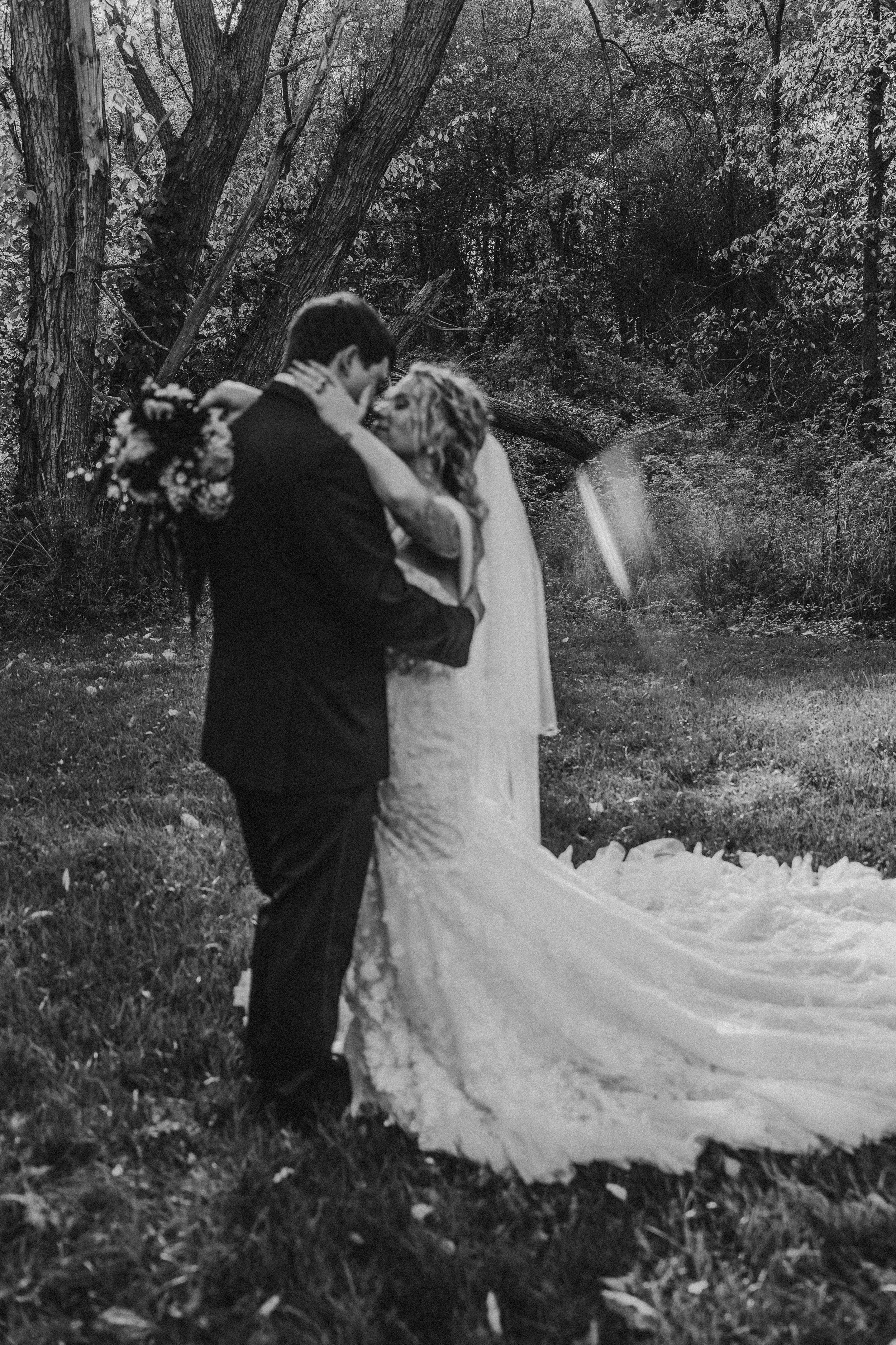 A black and white wedding photo shows a couple embracing in a romantic woodland setting with a trailing gown.