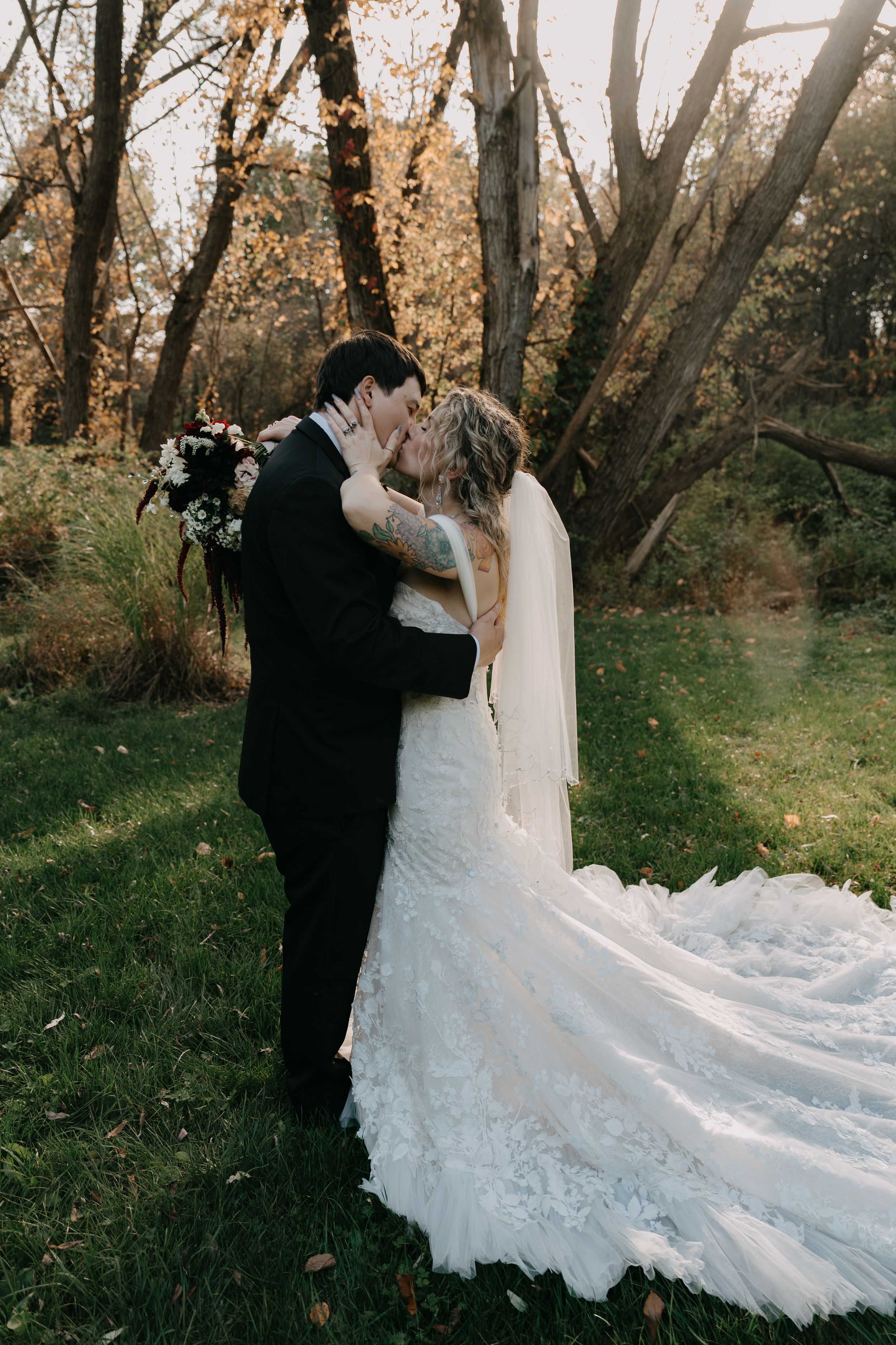Newlyweds share tender kiss in wooded setting with sunlight filtering through fall foliage.