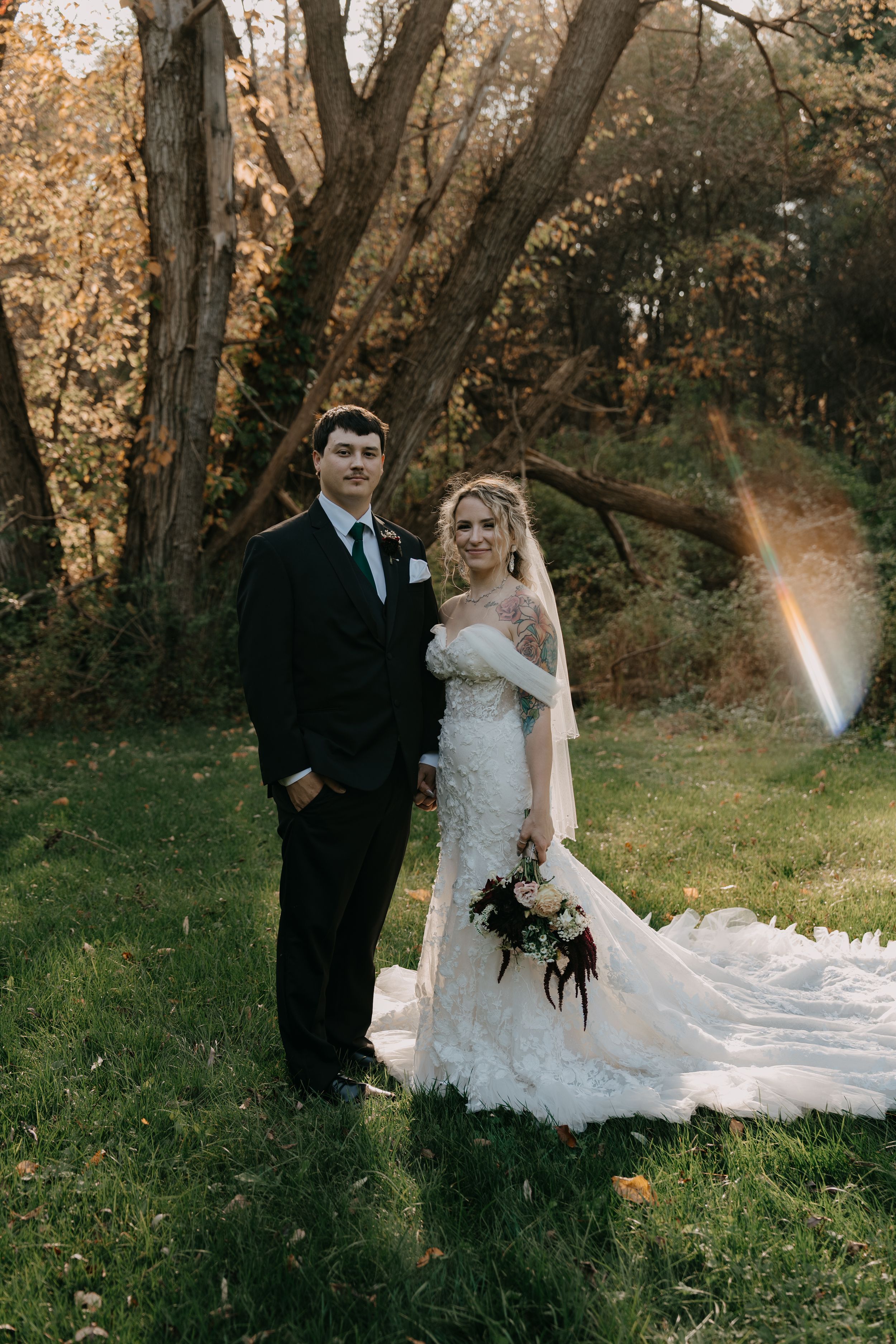 Newlyweds posing for a portrait in a wooded area with autumn foliage and golden sunlight.
