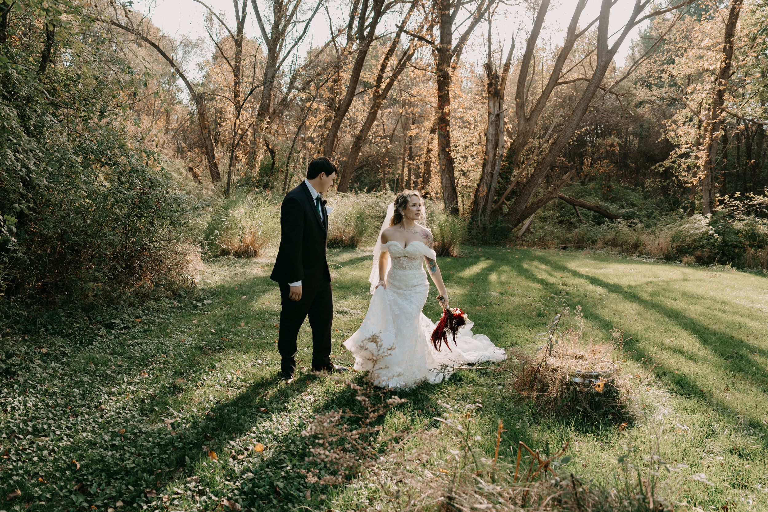 A couple walks through a sunlit woodland path, with the bride holding her wedding dress and bouquet in autumn scenery.