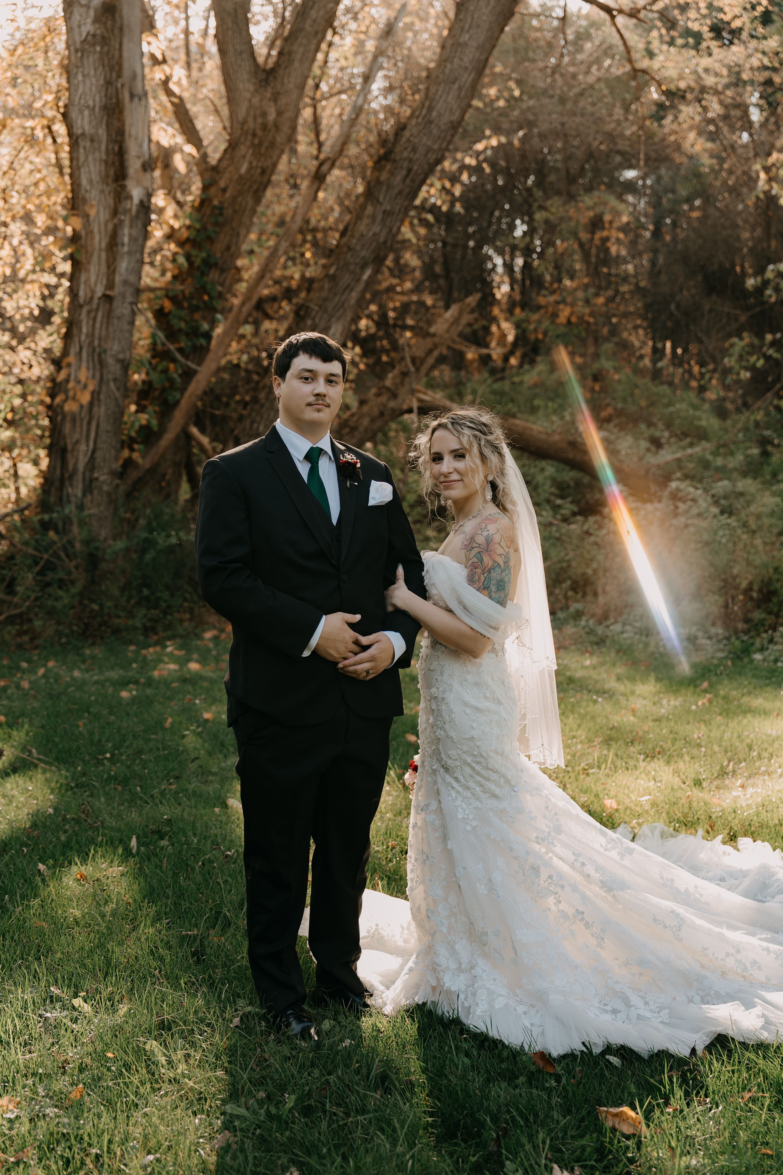 A bride in a lace wedding gown and groom in black suit pose together in a golden autumn forest setting.