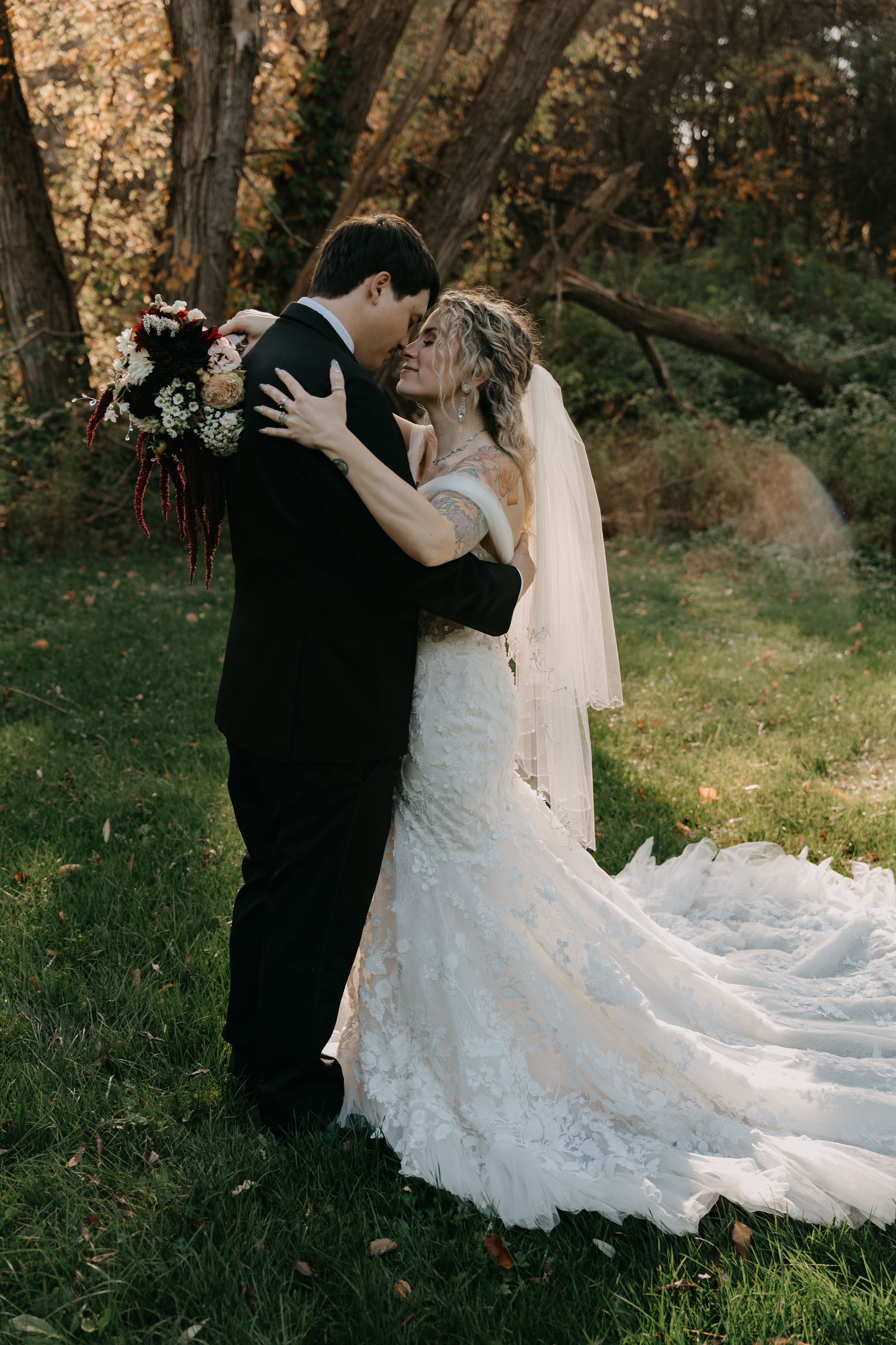 Romantic couple sharing intimate embrace in autumn forest with bride wearing flowing lace gown and delicate veil.