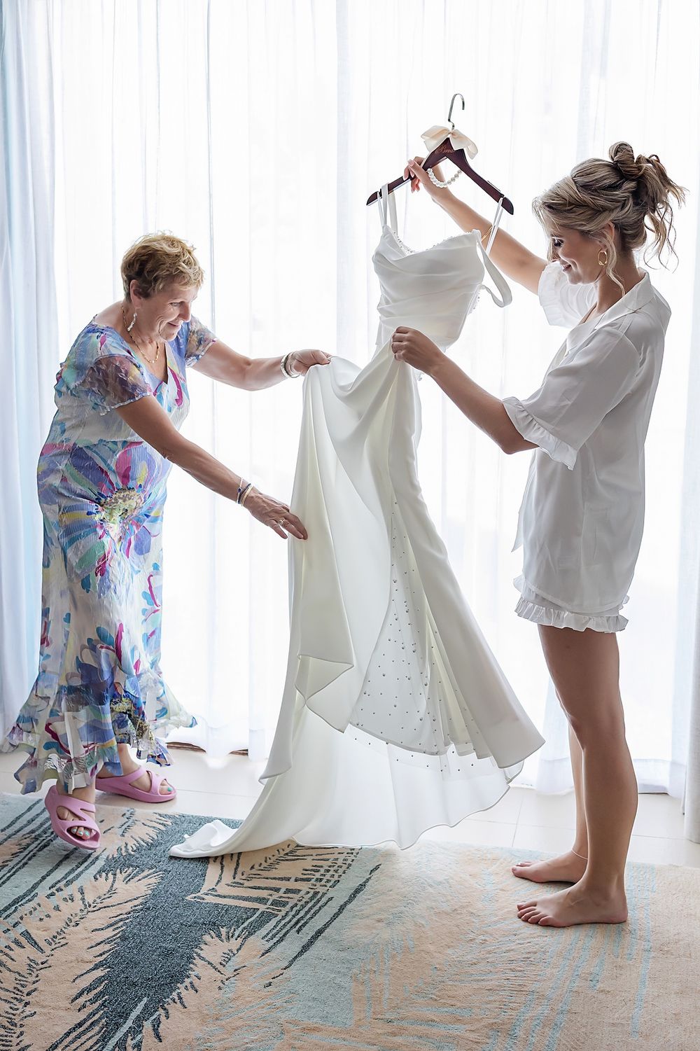 A woman in a floral dress and woman in white robe carefully examine a beautiful white wedding gown in a sunlit room.