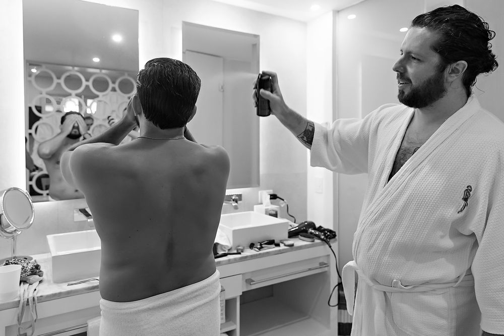 A hairdresser in a white robe uses hair tools while styling a customer in a backstage dressing room with mirrors.