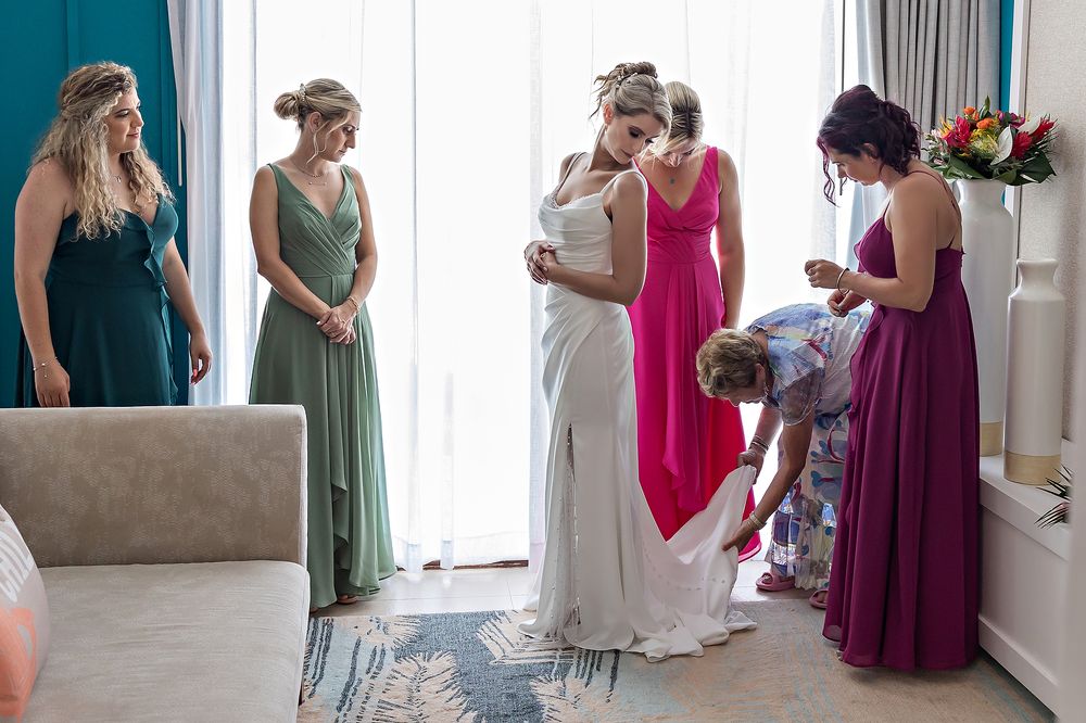 Group of women in elegant dresses of teal, green, pink and purple gather in a bright hotel room before an event.