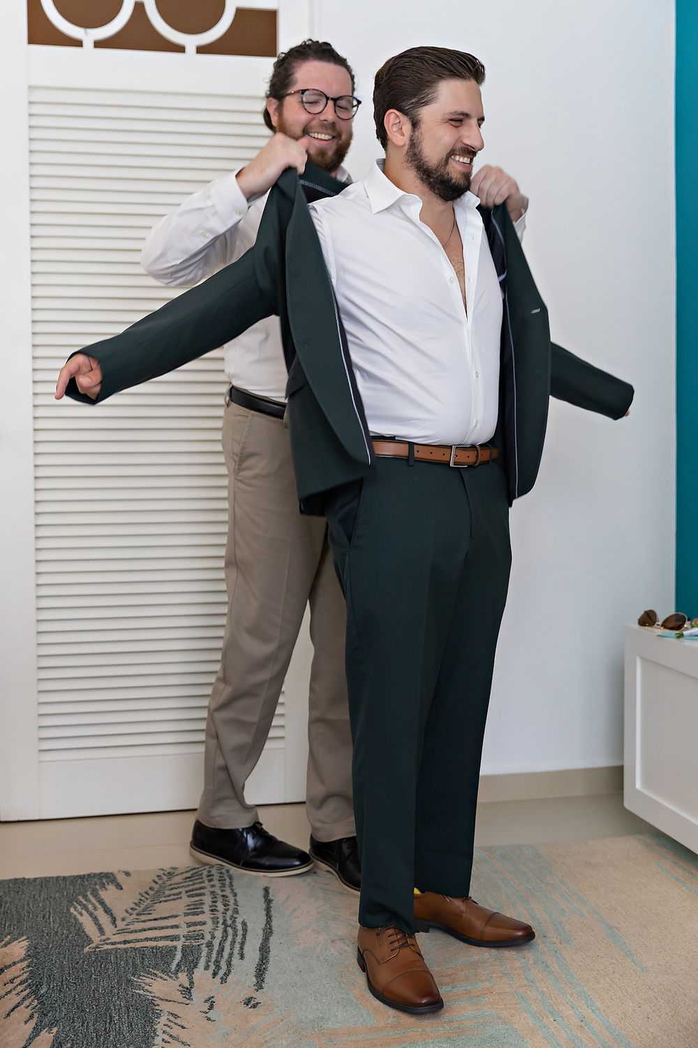Two men helping with formal attire fitting, one assisting the other with a dark suit jacket.