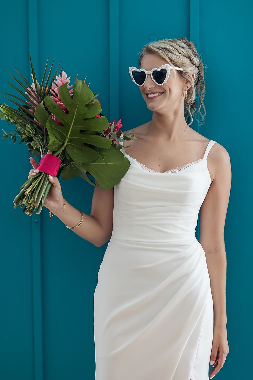 A person in a white wedding dress and sunglasses holds a tropical bouquet with pink flowers against a teal wall.