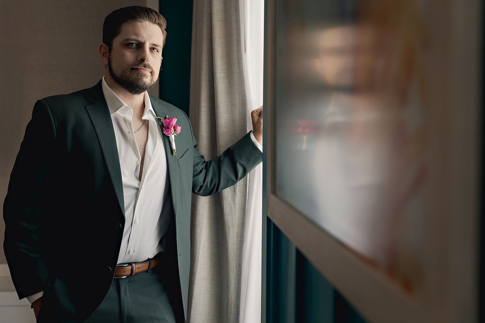 A groom in a dark suit and boutonniere stands thoughtfully by a window with natural lighting.