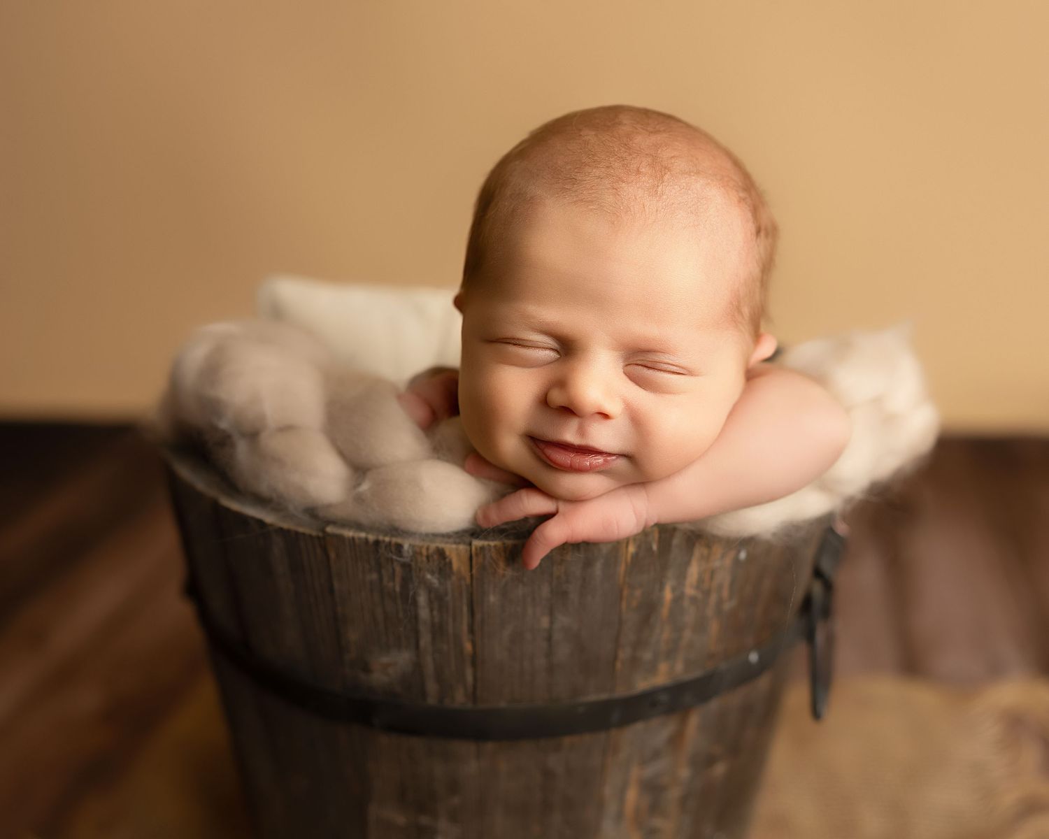 A peaceful sleeping newborn rests in a rustic wooden bucket lined with soft white fabric on a brown surface.