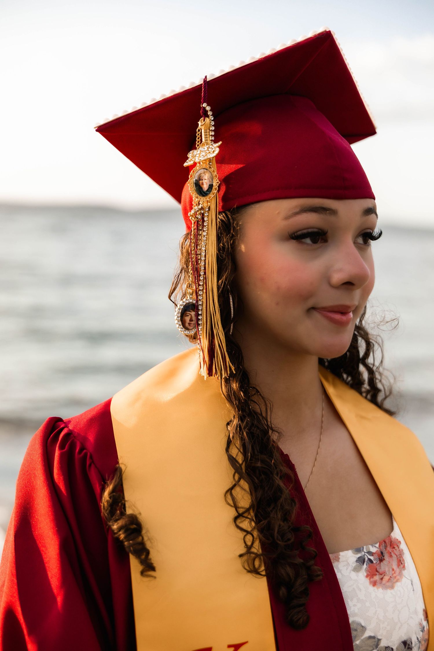 Graduate celebrates at sunset on sandy beach wearing floral dress and academic cap.