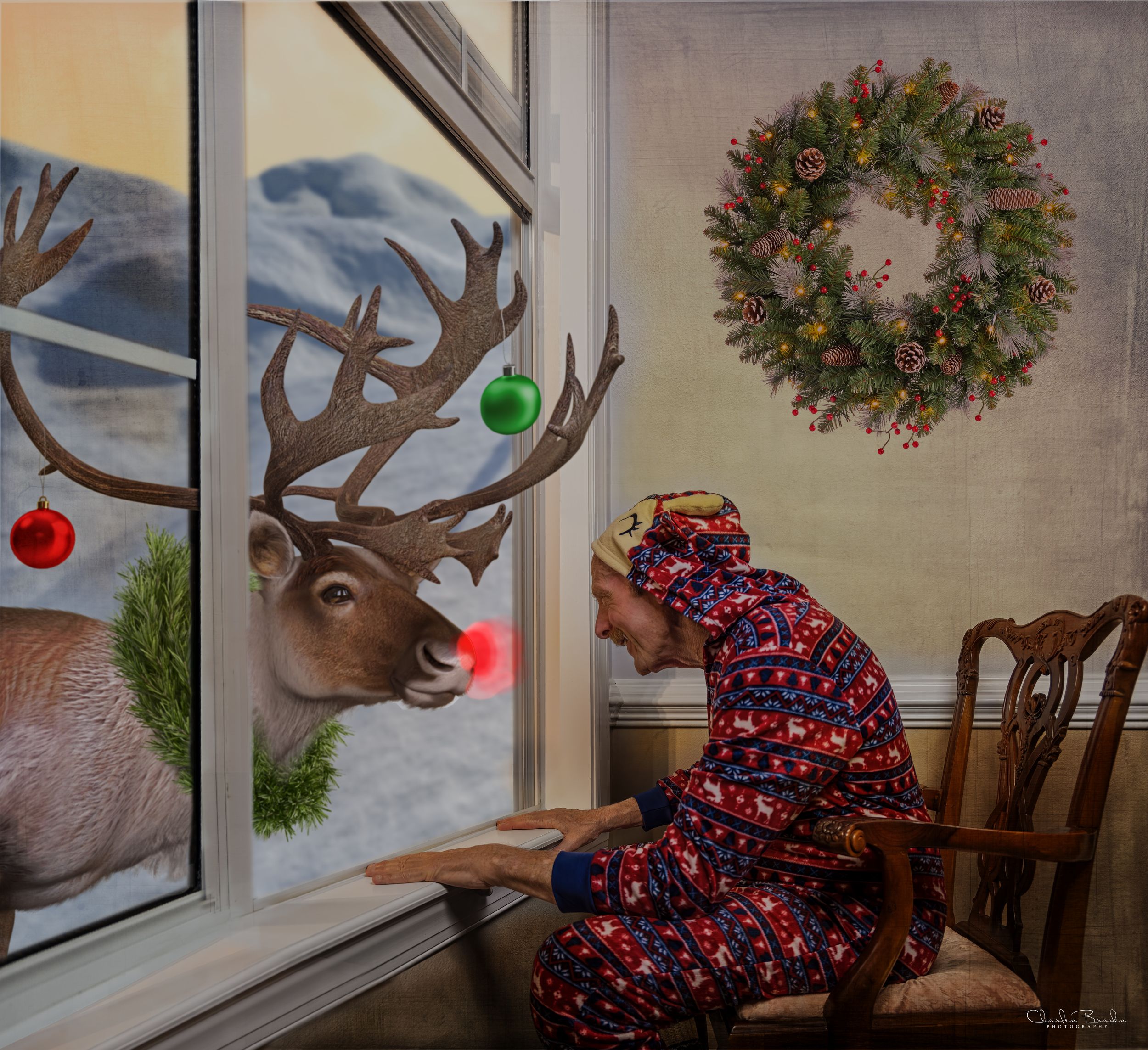 Person in holiday pajamas watches a reindeer decoration through window with Christmas wreath on wall.