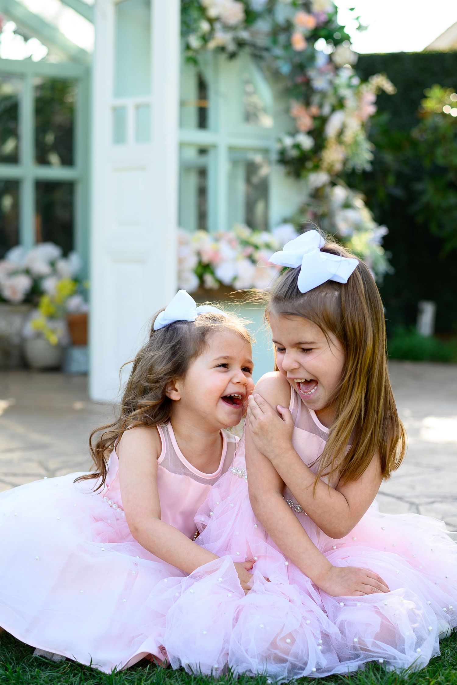 Two children in pink tulle dresses and white hair bows laugh and play together on the grass outside a house.