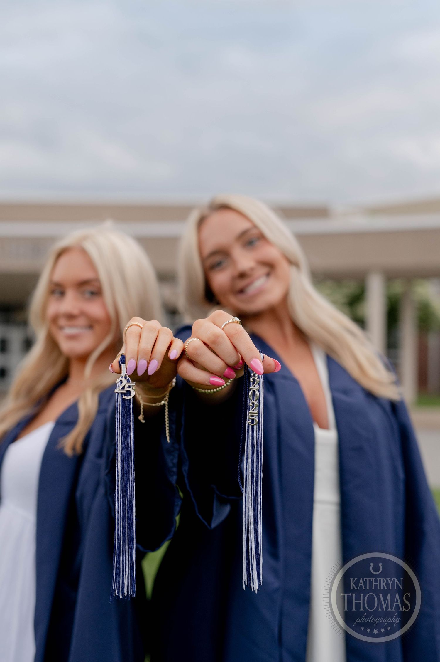 Two graduates in dark blue gowns display their silver tassels while celebrating their academic achievement.