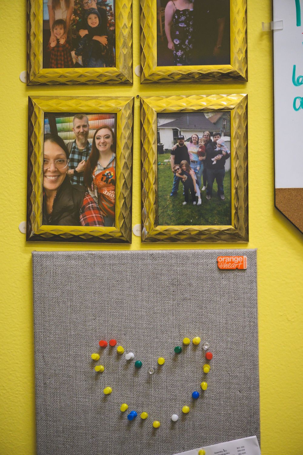 Photos displayed on a yellow wall with decorative gold frames and a gray bulletin board featuring a colorful pushpin heart design.