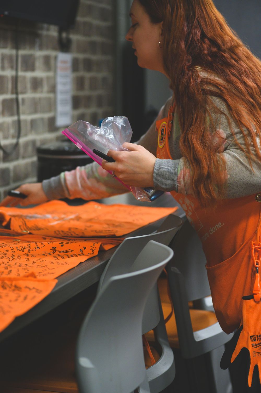 Person with long red hair wearing orange apron checks phone while sitting at table with orange t-shirts.