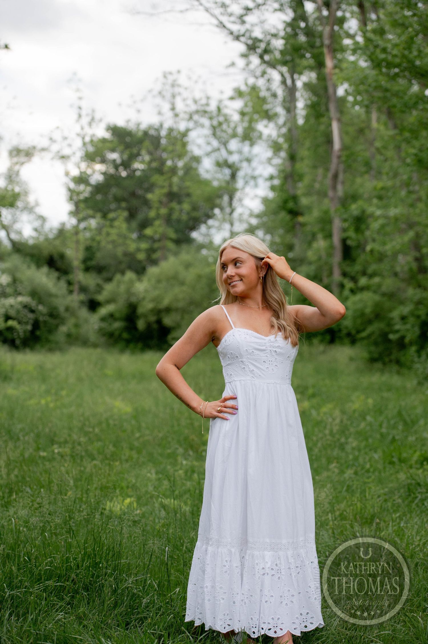 Young woman in flowing white summer maxi dress poses in a lush green meadow surrounded by trees.