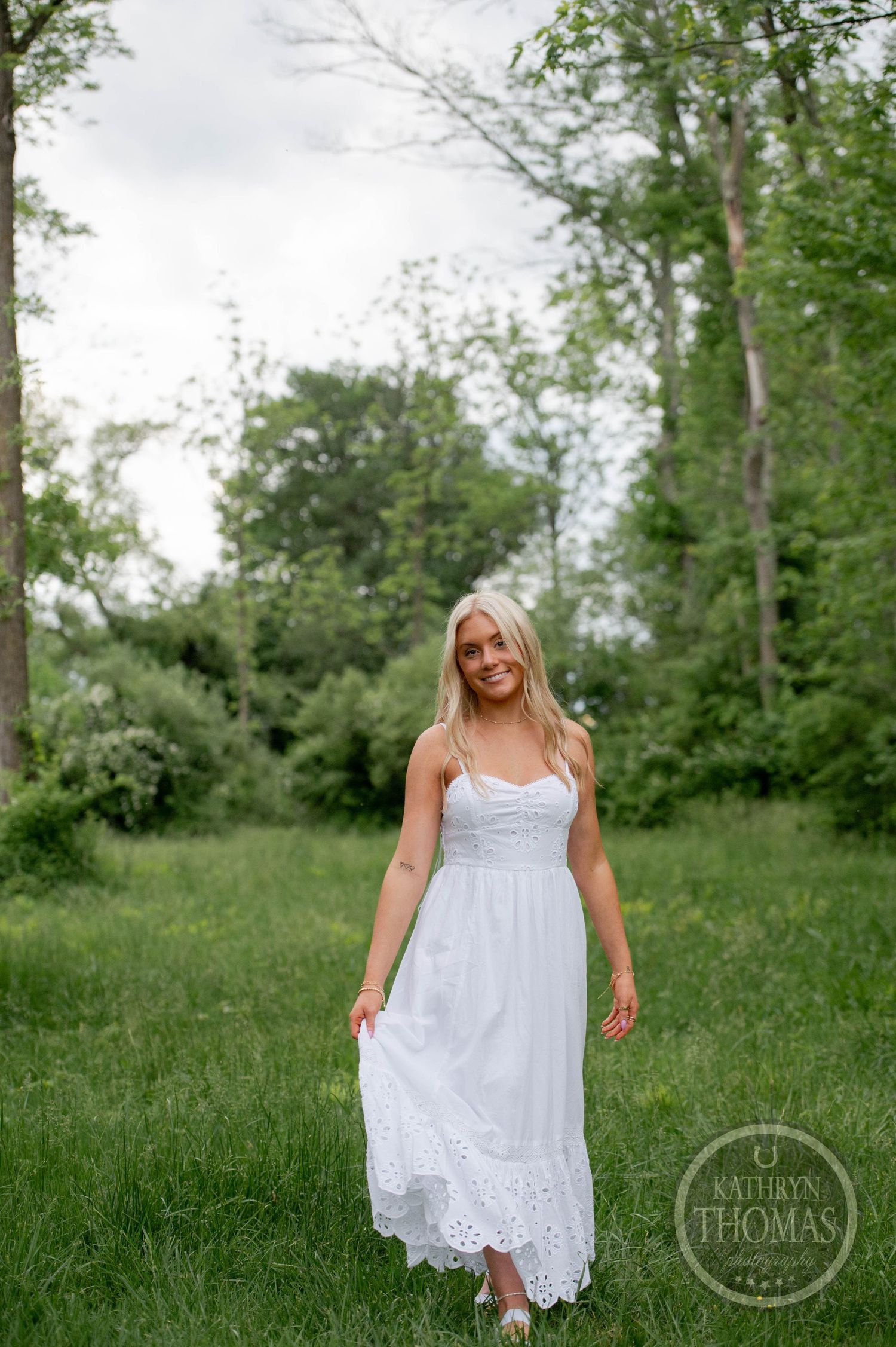 A woman in a flowing white sundress walks through a lush green summer meadow surrounded by trees.