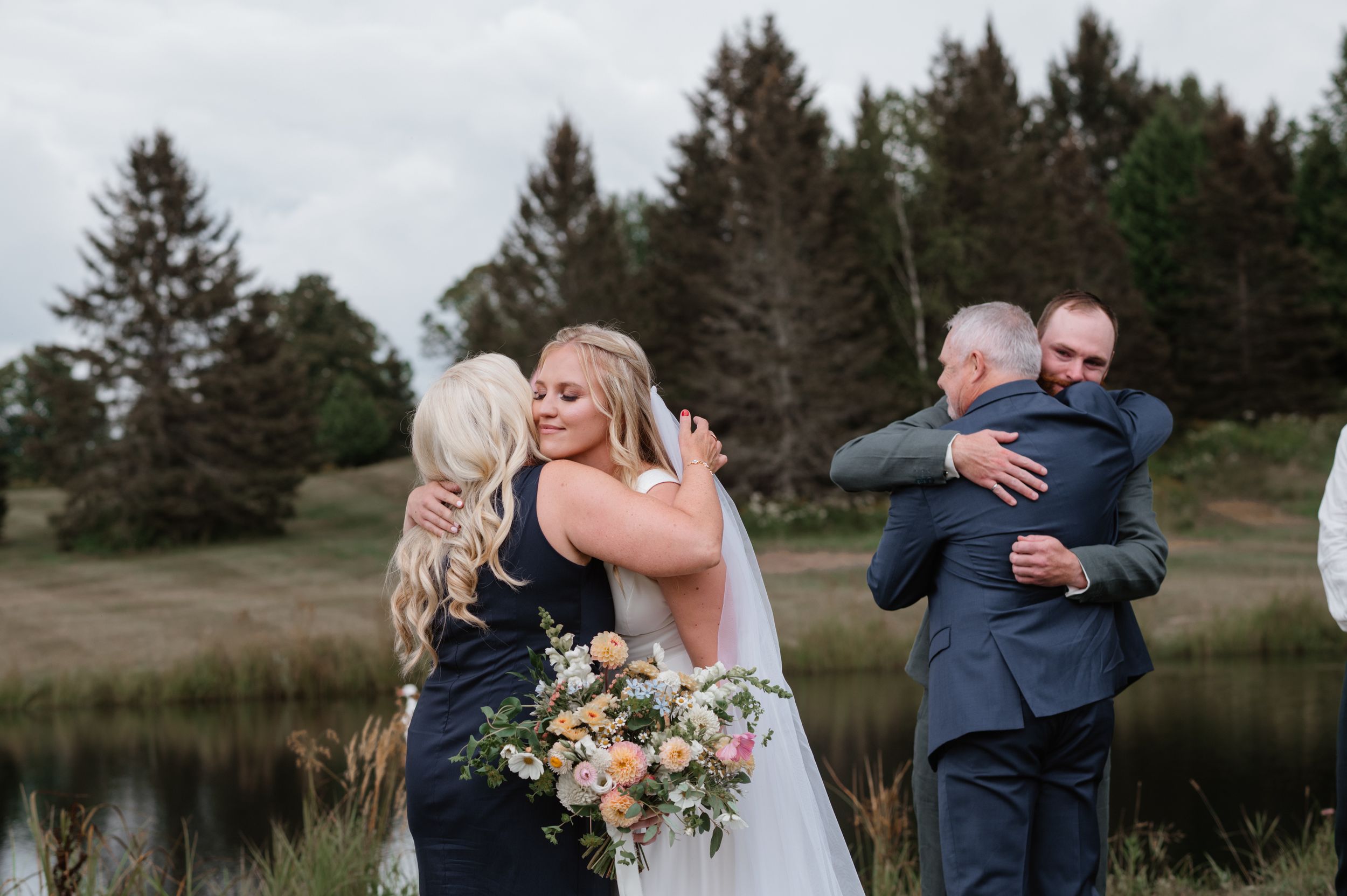 Wedding party members embrace and share emotional moments beside a pond with evergreen trees in the background.