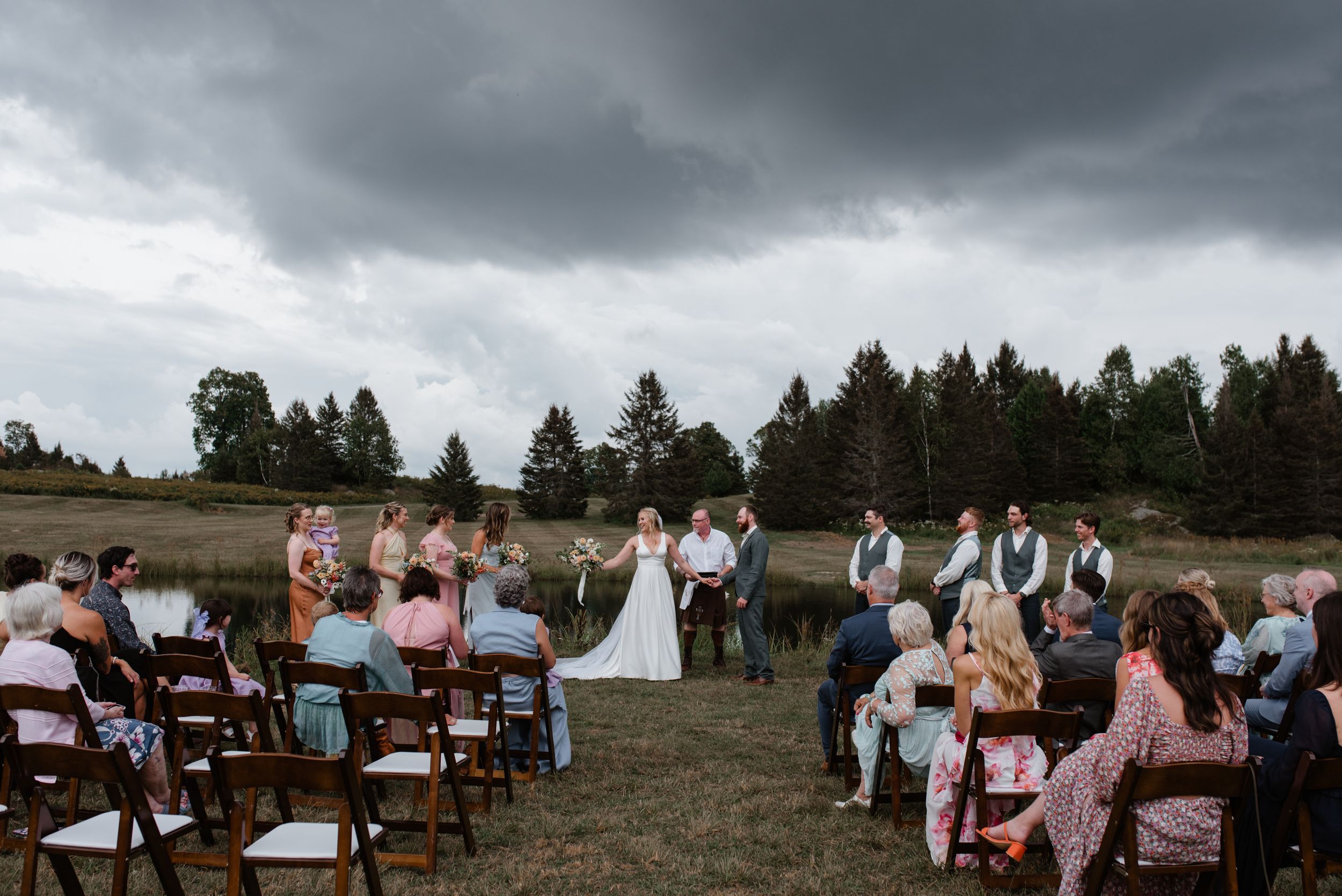 A rustic outdoor wedding ceremony surrounded by evergreen trees under moody storm clouds in a rural setting.