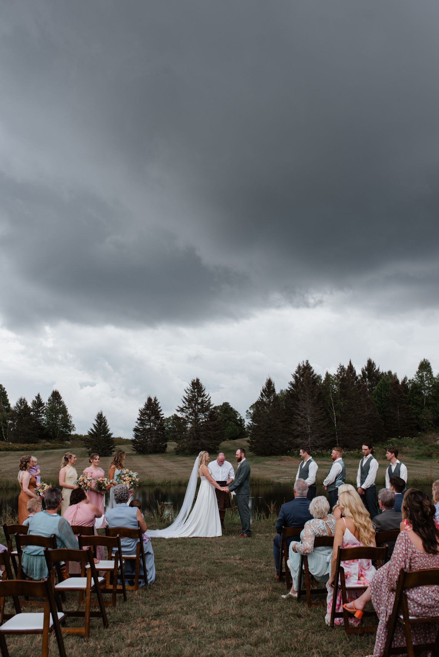An outdoor wedding ceremony in a rustic field with storm clouds gathering dramatically overhead in the background.