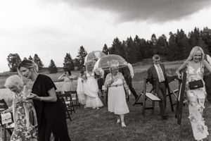 A wedding party stands on a grassy hillside as guests hold umbrellas during an outdoor celebration.