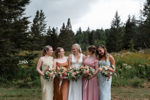 Wedding party in mix-matched pink and earth tone dresses stands together outdoors against mountain forest backdrop.