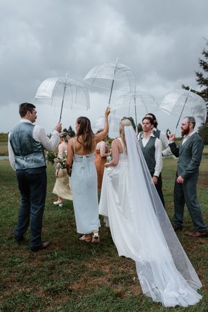 People holding clear umbrellas and wearing formal attire pose together outdoors during a wedding photoshoot.