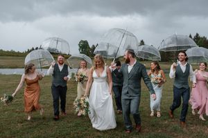 Wedding party running and jumping in a field together while holding umbrellas during a rainy celebration.