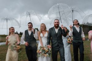 Wedding party laughs together while holding clear umbrellas on overcast day with bride holding colorful bouquet.