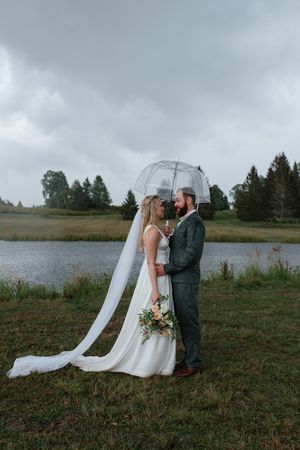 Two people embrace under a clear umbrella during a wedding photo session by a lake on an overcast day.