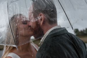 Two people share an intimate moment under a clear umbrella in the rain during what appears to be a wedding day.